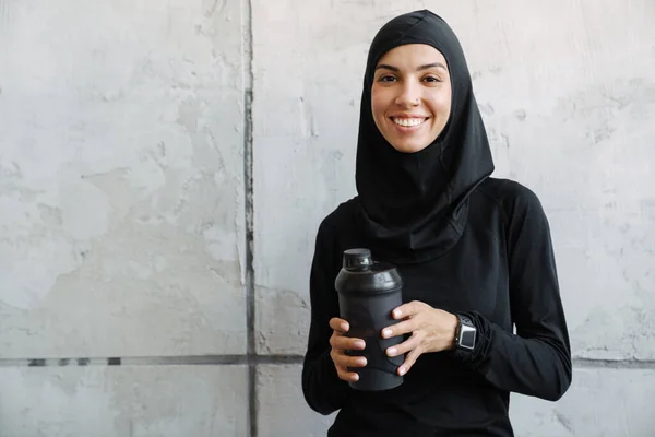 年轻的穆斯林妇女头戴头巾站在屋里笑着 手里拿着水瓶 — 图库照片