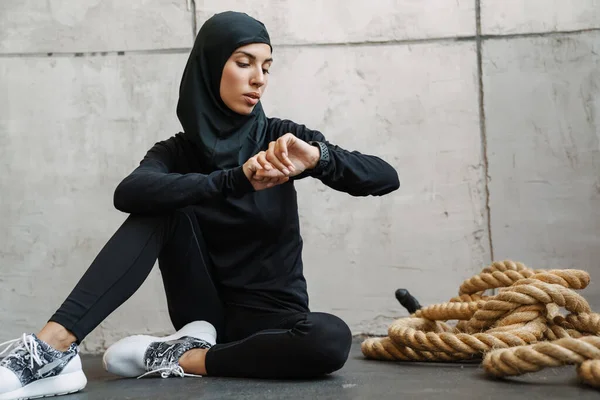 Młoda Muzułmanka Hidżabie Patrzy Smartwatch Siedząc Podłodze Domu — Zdjęcie stockowe