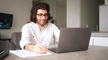Beyaz gömlek ve gözlük takan yakışıklı bir Türk dairede dizüstü bilgisayarıyla notlar alıyor.