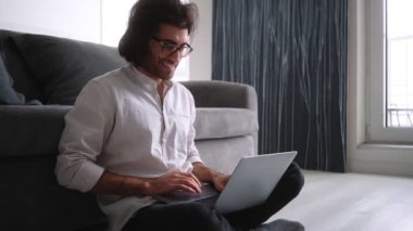 Beyaz gömlek ve gözlük takan gülümseyen bir Türk, laptopuyla içeride oturuyor.