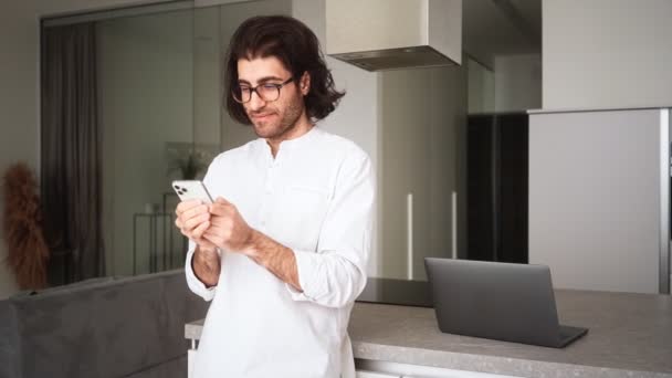 一位身穿白衬衫 戴眼镜 面带微笑的乌黑头发男子站在公寓里用手机 — 图库视频影像