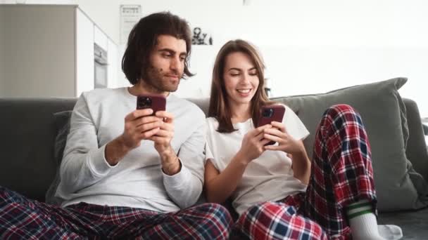 笑顔のカップルの女性と男はソファの上に座っているソーシャルメディアのコンテンツを探しています屋内 — ストック動画