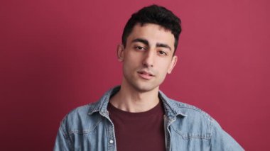 Esmer bir adam kameraya bakarken esniyor. Stüdyonun kırmızı arka planında tek başına duruyor.