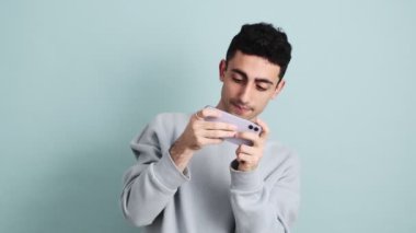 Mutlu bir esmer adam cep telefonuyla oyun oynuyor. Stüdyonun mavi arka planında izole bir şekilde duruyor.