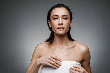 Havlu giymiş beyaz kadın gri arka planda yalnız başına poz veriyor ve kameraya bakıyor.