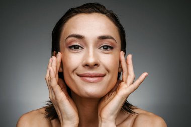 Yarı çıplak bir kadın yüzüne dokunuyor ve gri arka planda izole edilmiş kameraya gülümsüyor.