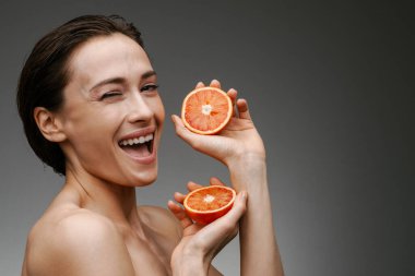 Sağlıklı orta yaşlı, üstsüz bir kadın. Gri arka planda greyfurt tutuyor, göz kırpıyor.