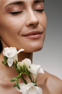 Gri arkaplanda beyaz çiçekle poz veren Avrupalı üstsüz orta kadın