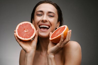 Gri arka planda izole edilmiş greyfurtla poz veren beyaz tişörtsüz kadın gülüyor.