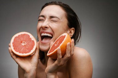 Gri arka planda izole edilmiş greyfurtla poz veren beyaz tişörtsüz kadın gülüyor.