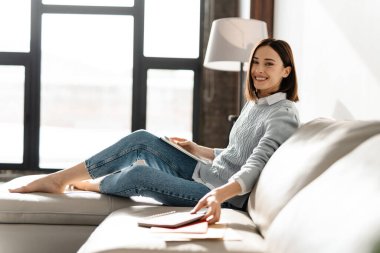 Evdeki kanepede otururken tablet bilgisayar kullanan gülümseyen genç kadın.