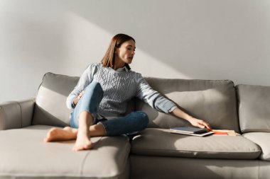 Evdeki kanepede otururken elinde defterler tutan beyaz bir kadın.