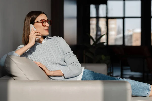 Gözlüklü Genç Beyaz Kadın Evdeki Koltukta Otururken Cep Telefonuyla Konuşuyor — Stok fotoğraf