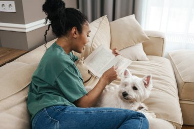 Orta yaşlı Afrikalı bir kadın evinde köpeğiyle kanepede oturuyor. Kitap okumak