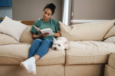 Evdeki kanepede köpeğiyle otururken kitap okuyan genç siyahi kadın.