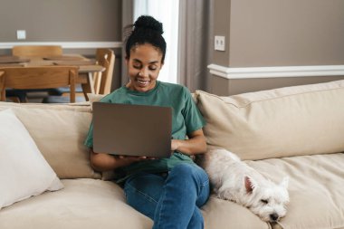 Evdeki kanepede köpeğiyle otururken gülümseyen siyah kadın dizüstü bilgisayar kullanıyor.