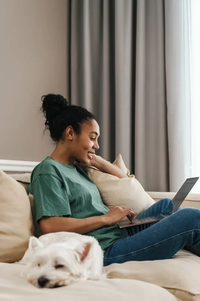 Smiling black woman using laptop while sitting with her dog on sofa at home
