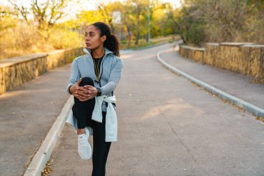 Odaklanmış Afrikalı Amerikalı sporcu kadın parkta çalışırken bacağını esnetiyor.
