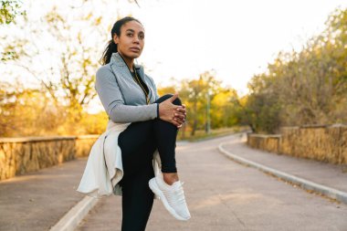 Odaklanmış Afrikalı Amerikalı sporcu kadın parkta çalışırken bacağını esnetiyor.