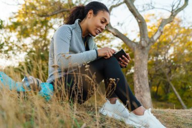 Gülümseyen Afro-Amerikan sporcu kadın parkta otururken cep telefonu kullanıyor.