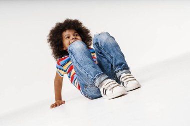 Sırıtan küçük Afrikalı çocuk, günlük kıyafetlerini giymiş, duvarın arkasında, yerde oturuyor.