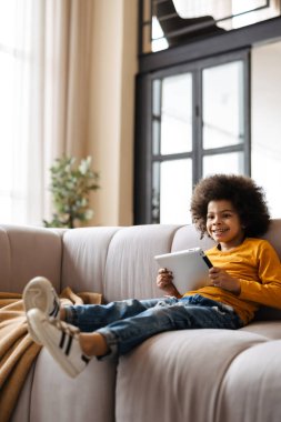 Siyah kıvırcık çocuk evde kanepede otururken gülümsüyor ve tablet bilgisayar kullanıyor.