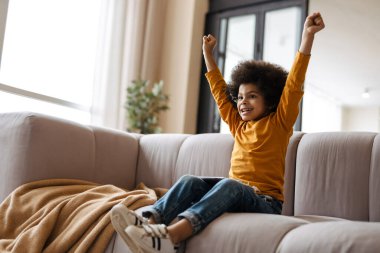 Siyah kıvırcık çocuk evde kanepede otururken el kol hareketi yapıyor ve tablet bilgisayar kullanıyor.