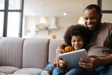 Siyah baba ve oğul evde kanepede otururken gülümsüyor ve tablet bilgisayar kullanıyor.