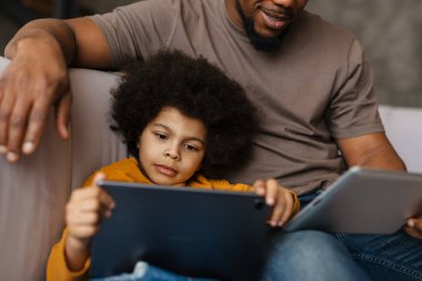 Siyah baba ve oğlu evdeki kanepede otururken tablet bilgisayar kullanıyorlar.