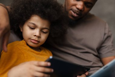 Siyah baba ve oğlu evdeki kanepede otururken tablet bilgisayar kullanıyorlar.