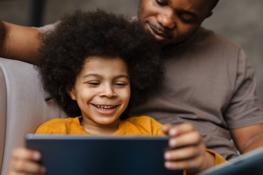 Siyah baba ve oğul evde kanepede otururken gülümsüyor ve tablet bilgisayar kullanıyor.