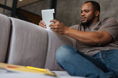 Siyah traşsız adam evdeki koltukta otururken tablet bilgisayar kullanıyor.