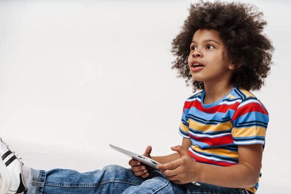 Preschooler african boy wearing t-shirt sitting over white wall, holding digital tablet computer, looking aside