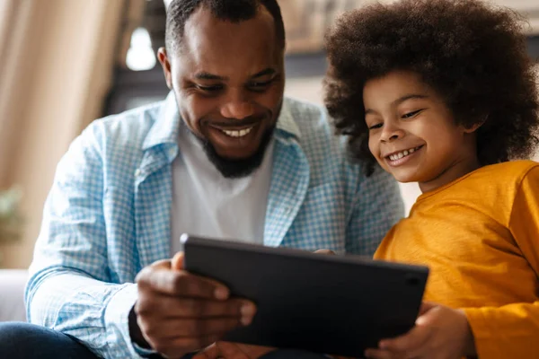 Black father and son smiling and using tablet computer while sitting on sofa at home