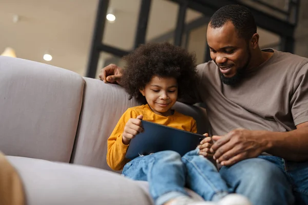 Black father and son smiling and using tablet computer while sitting on sofa at home