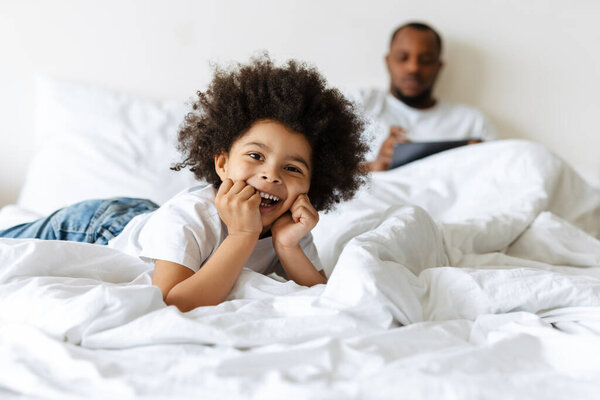 Black boy laughing while his father using tablet computer in bed at home