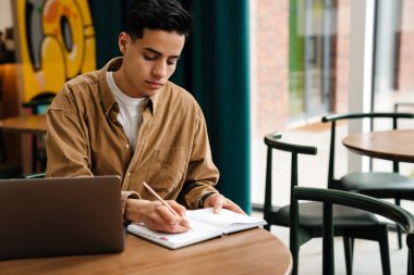 Kafe masasında dizüstü bilgisayarla oturan genç, sakin İspanyol öğrenci notlar alıyor.