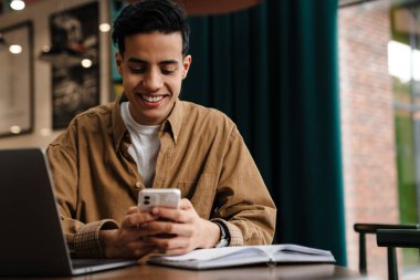 Kafe masasında dizüstü bilgisayarla oturan ve elinde cep telefonu olan gülümseyen genç İspanyol öğrenci.