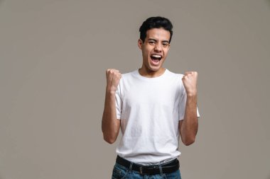 Gülümseyen esmer tişörtlü genç adam gri duvar arkasında duruyor, başarıyı kutluyor.