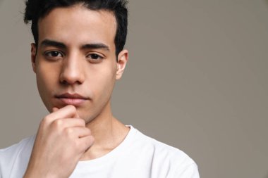 Tişörtlü dalgın, esmer bir adam gri duvar arkasında kameraya bakıyor.