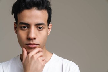 Tişörtlü dalgın, esmer bir adam gri duvar arkasında kameraya bakıyor.