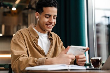 Kafe masasında dizüstü bilgisayarla oturan gülümseyen genç İspanyol öğrenci elinde cep telefonu, video izliyor.