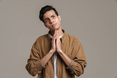 Esmer, İspanyol bir adam avuç içlerini bir arada tutuyor ve gri arka planda yalnız bir şekilde yukarı bakıyor.