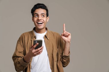 Gülümseyen esmer tişörtlü genç adam gri duvar arkasında duruyor, cep telefonunu tutuyor, parmağıyla işaret ediyor.