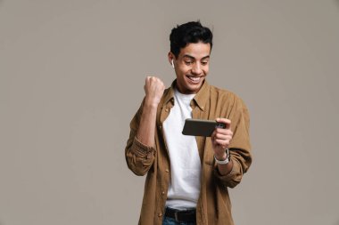 Gülümseyen, tişörtlü esmer adam gri duvar arkasında duruyor, cep telefonu tutuyor, oyunlar oynuyor, başarıyı kutluyor.