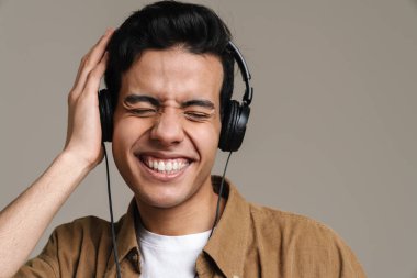 Gülümseyen, tişörtlü, gri duvar arkasında duran, kulaklıkla müzik dinleyen esmer bir adam.