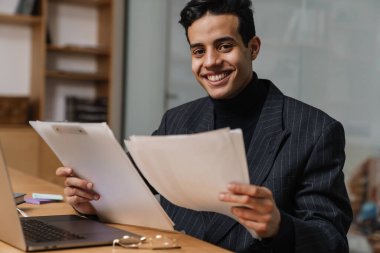 Orta yaşlı, esmer, takım elbiseli bir girişimci ofiste çalışıyor, masada dizüstü bilgisayar ve belgelerle oturuyor, evrak işlerine bakıyor.