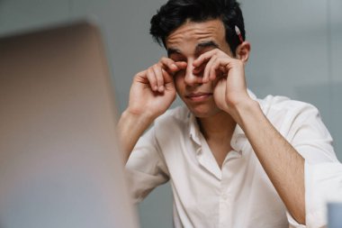 İş yerinde oturan, beyaz gömlekli, orta yaşlı esmer işadamı gözlerini ovuşturuyor.