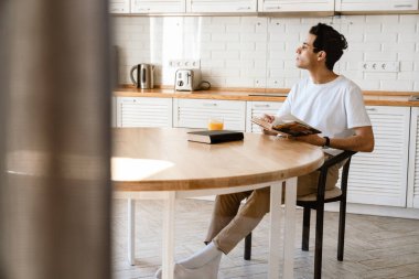 Genç İspanyol adam evde mutfakta kahvaltı ederken kitap okuyor, masada oturuyor.
