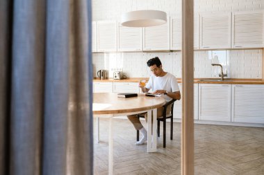Genç İspanyol adam evde mutfakta kahvaltı ederken kitap okuyor, masada oturuyor.